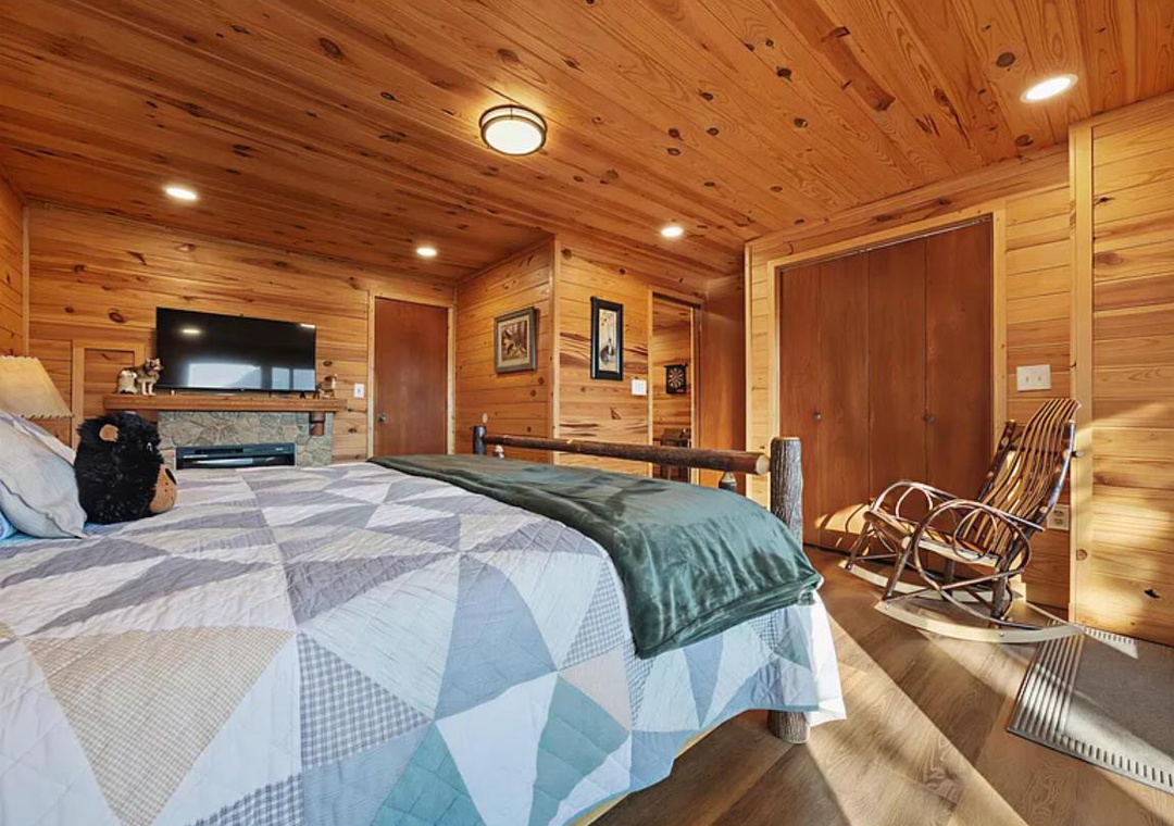 Wrap yourself in the warmth of knotty pine walls and soft quilts in this inviting bedroom, where a cozy fireplace and rustic rocking chair promise restful mountain evenings.