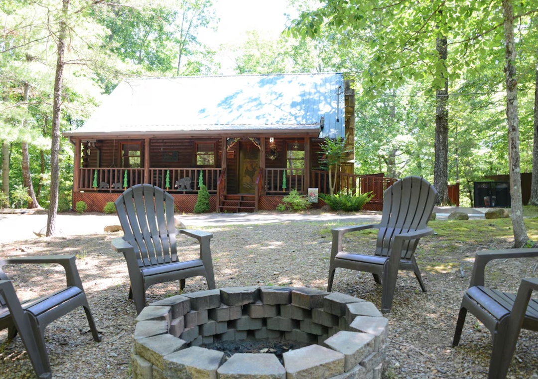 Charming forest cabin with welcoming covered porch and stone fire pit area surrounded by mature trees and peaceful woodland setting.