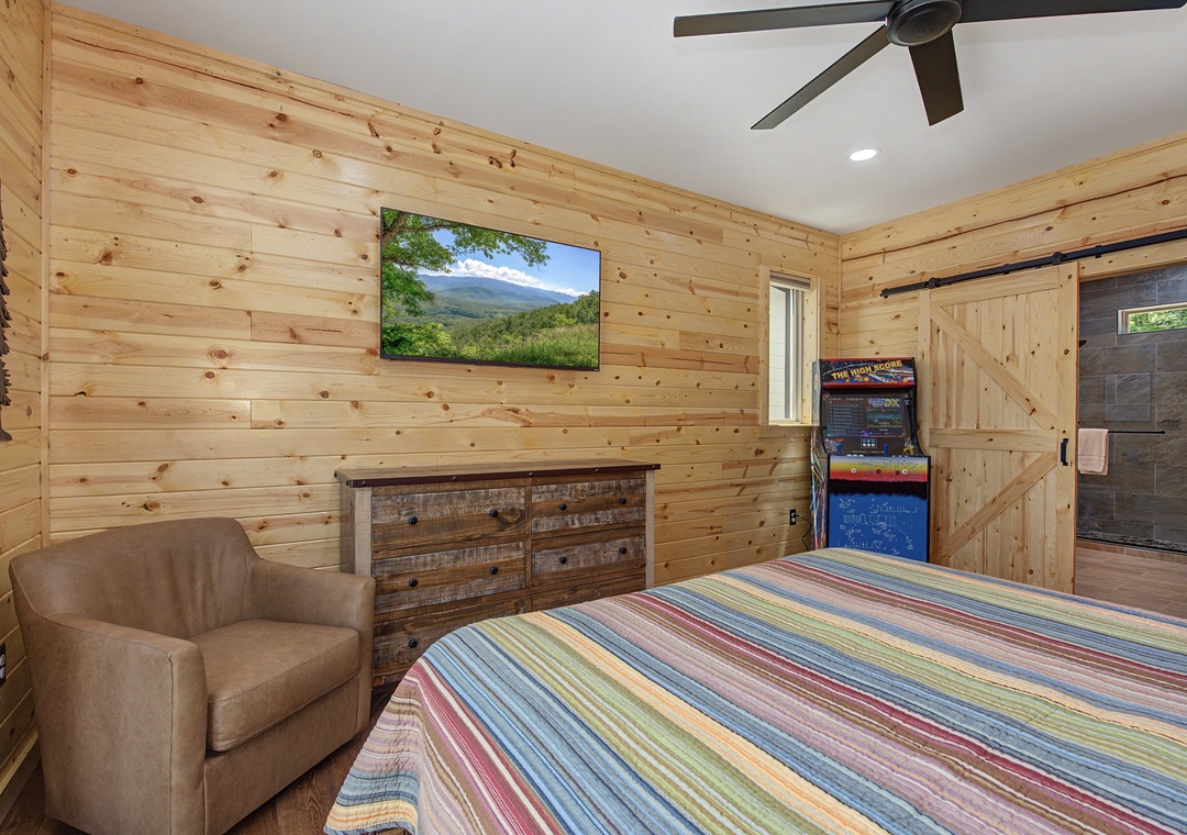 Your cozy cabin bedroom features warm pine walls, a comfortable chair, entertainment options including a TV and arcade game, plus mountain views and an ensuite bathroom for your convenience.