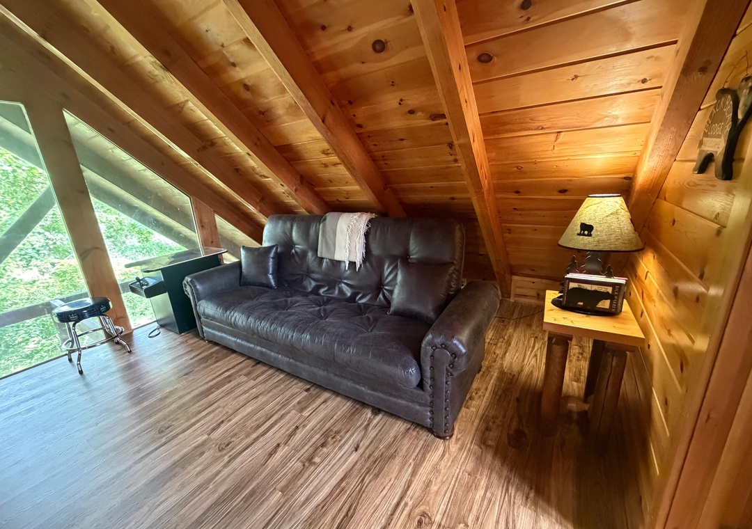 Cozy up in this charming loft living space with vaulted wood ceilings, a plush leather sofa, and natural light streaming through large windows overlooking lush greenery—your perfect retreat awaits.