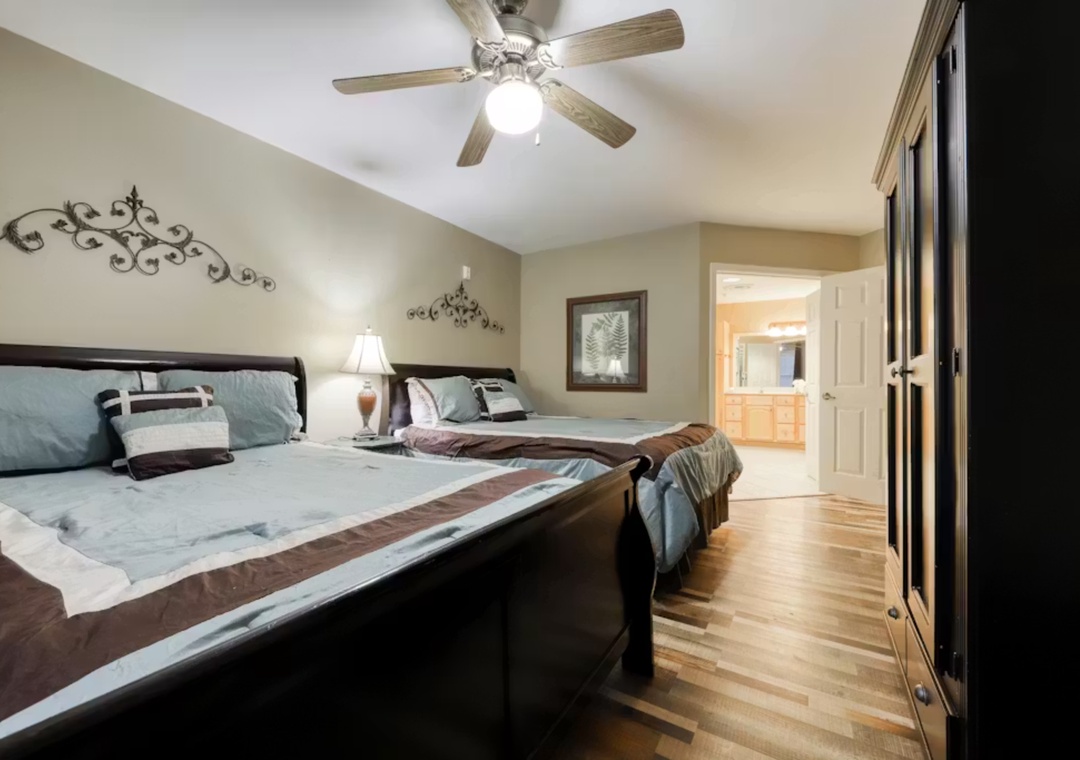 Rest peacefully in this elegant bedroom featuring two comfortable beds with soft blue linens, warm hardwood floors, and decorative ceiling fan.
