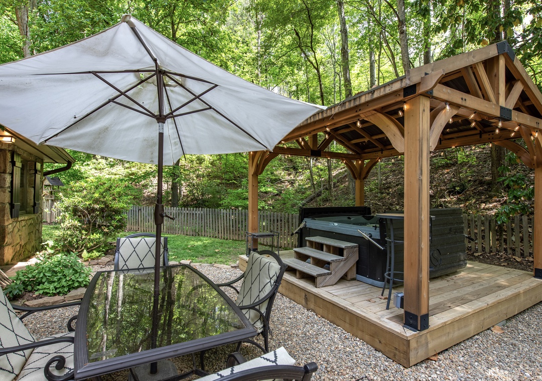 Unwind in your private wooded retreat with a soothing hot tub nestled under a timber pavilion, surrounded by lush greenery and tranquil outdoor seating.