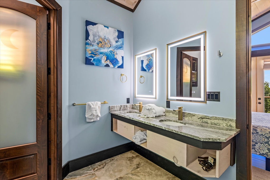 Picture yourself freshening up at this stunning double vanity with elegant marble countertops and warm brass fixtures. The soft blue walls and glowing backlit mirrors create a spa-like ambiance that invites relaxation.