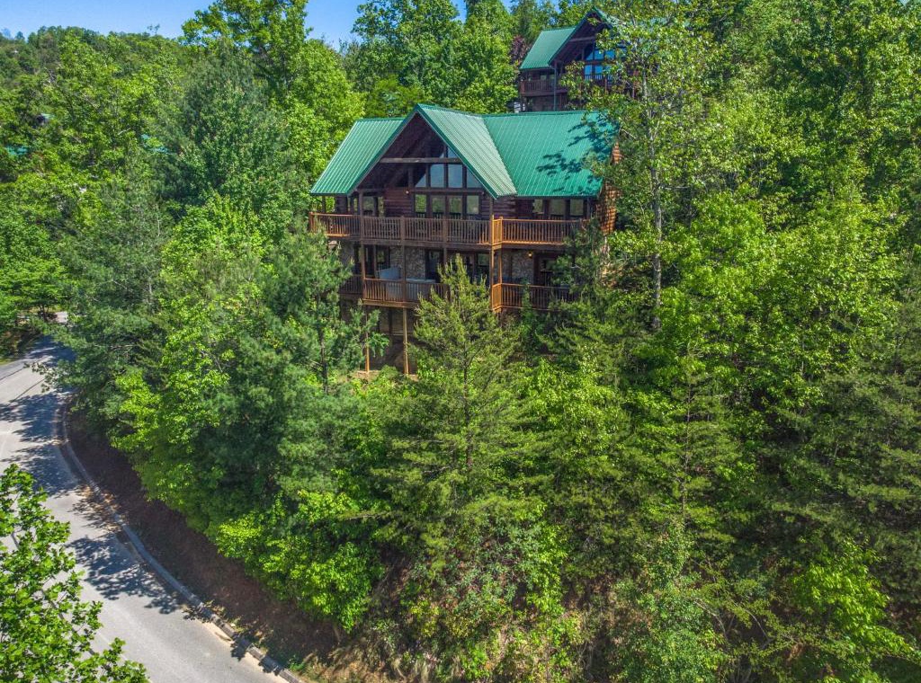 Rustic log cabin with green metal roof nestled among lush forest canopy, featuring multiple decks and mountain retreat architecture.