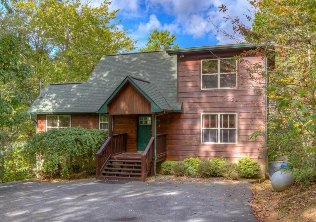A charming two-story wood cabin nestled in a wooded area, featuring a welcoming front entrance, private parking, and natural landscaping with mature trees providing peaceful seclusion.