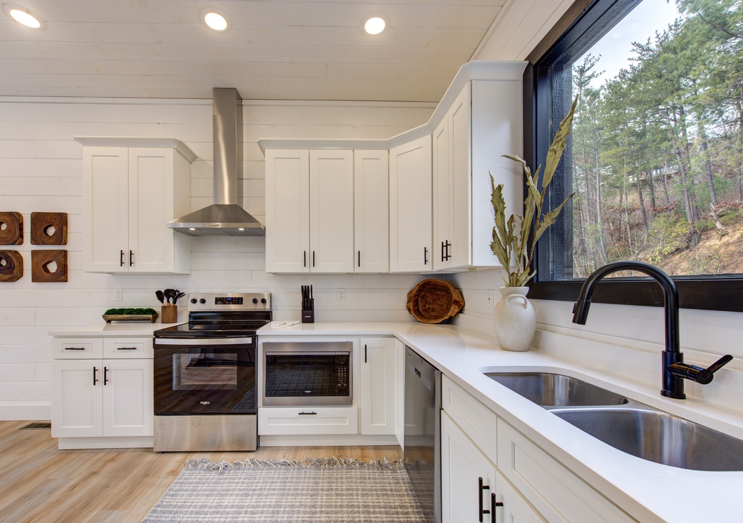 Your fully equipped kitchen features gleaming countertops, modern appliances, and natural forest views through the large window.