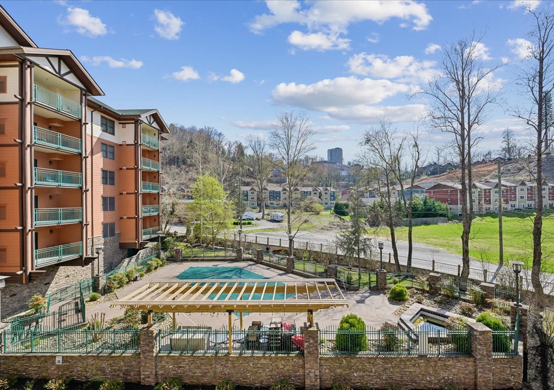 Spacious resort community featuring multiple buildings, outdoor pool with deck seating, green lawns, and city views in the distance.