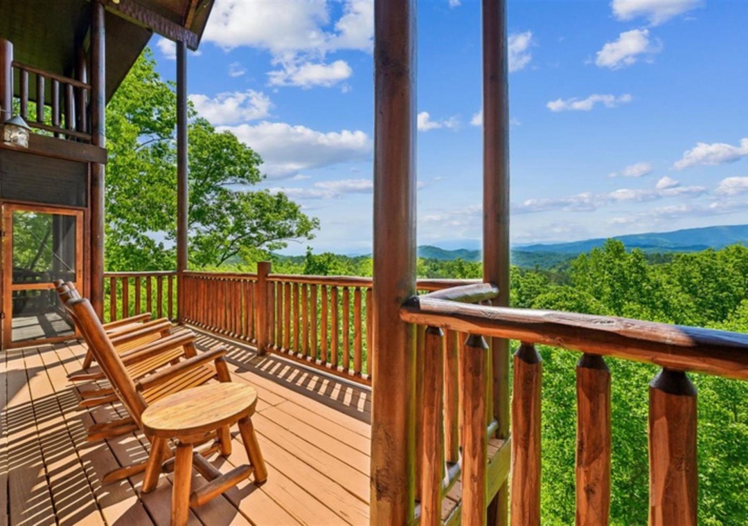 Expansive mountain vistas stretch across the horizon from this elevated wooden deck, surrounded by lush forest canopy. The covered balcony features classic rocking chairs positioned to take in the sweeping panoramic views of rolling blue ridges.