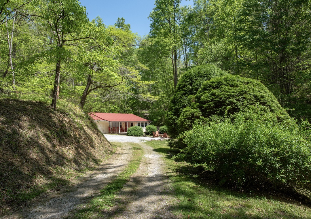A charming red-roofed retreat nestled in lush forest, accessible via a peaceful gravel path surrounded by vibrant spring greenery.