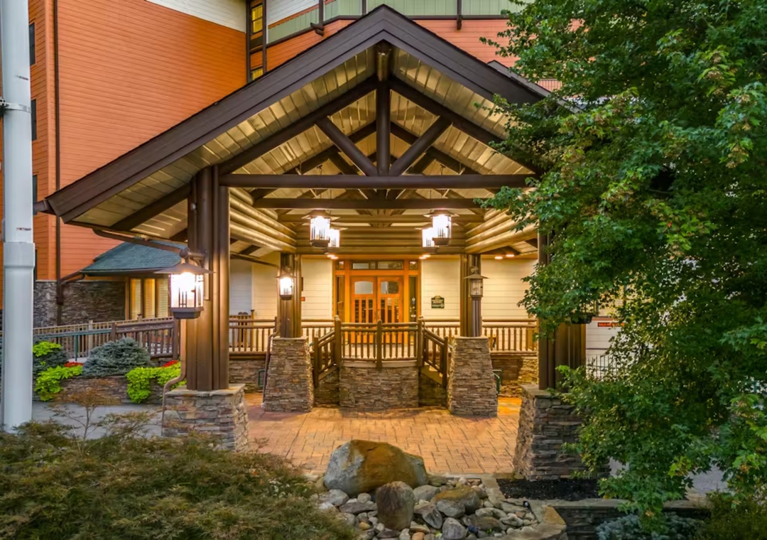 Elegant property entrance features warm wood beams, stone pillars, and welcoming lighting surrounded by mature landscaping.