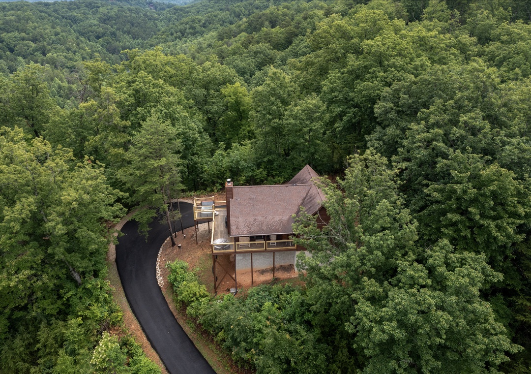 Aerial view of a private mountain cabin nestled within lush forest canopy, accessed by a secluded driveway and surrounded by natural woodland landscape.