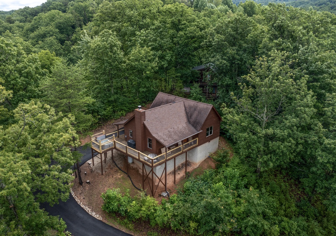Aerial view of a secluded cabin retreat nestled in lush mountain forest, featuring elevated decks and a scenic wooded hillside backdrop.