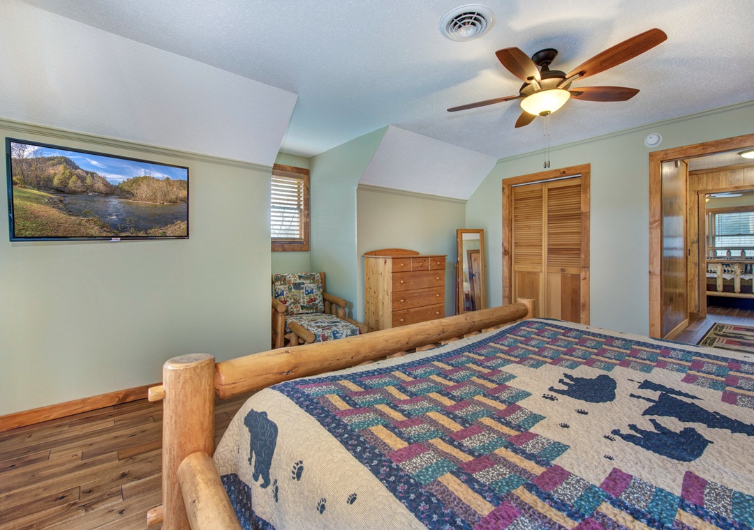 Relax in your tranquil bedroom with rustic log bed frame, cozy armchair, and modern amenities like ceiling fan and flat-screen TV. Warm wood tones and soft lighting create the perfect retreat.