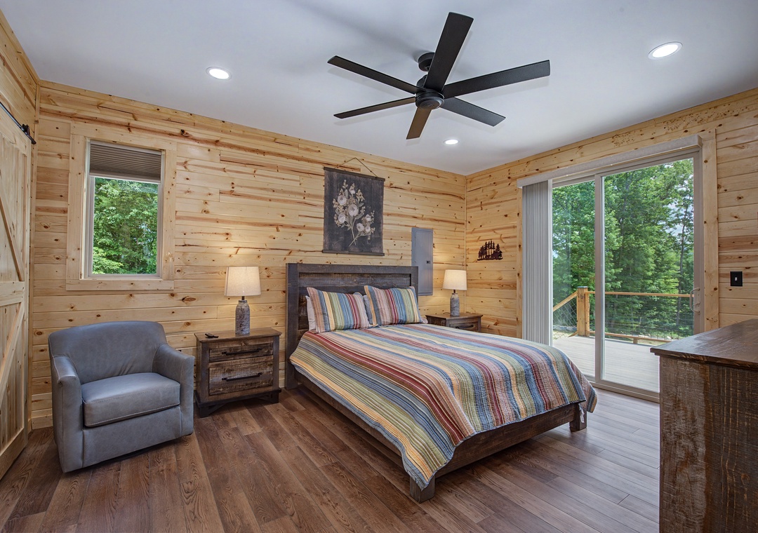 Step into your serene bedroom retreat with warm wood-paneled walls, a plush bed dressed in vibrant linens, and sliding glass doors opening to your private balcony surrounded by lush forest views.