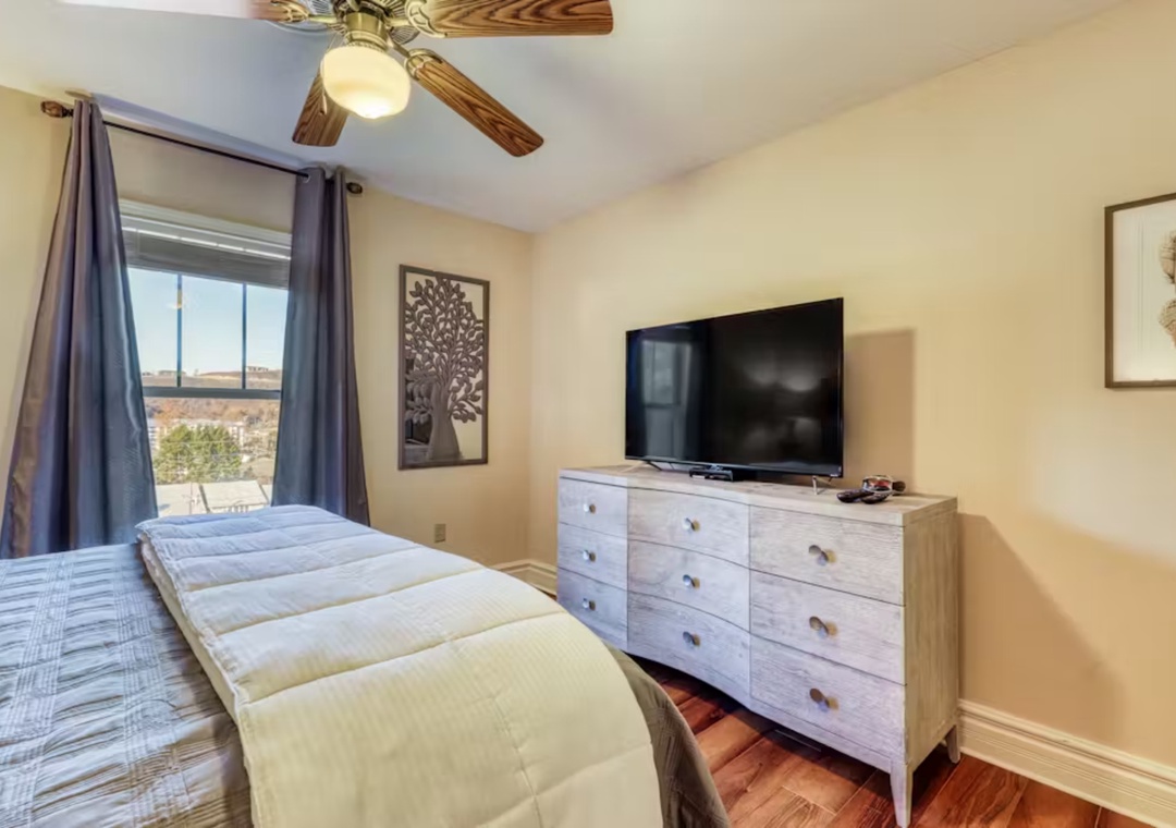 Unwind in your cozy bedroom with plush bedding, smart TV entertainment, and peaceful views through blue curtains.