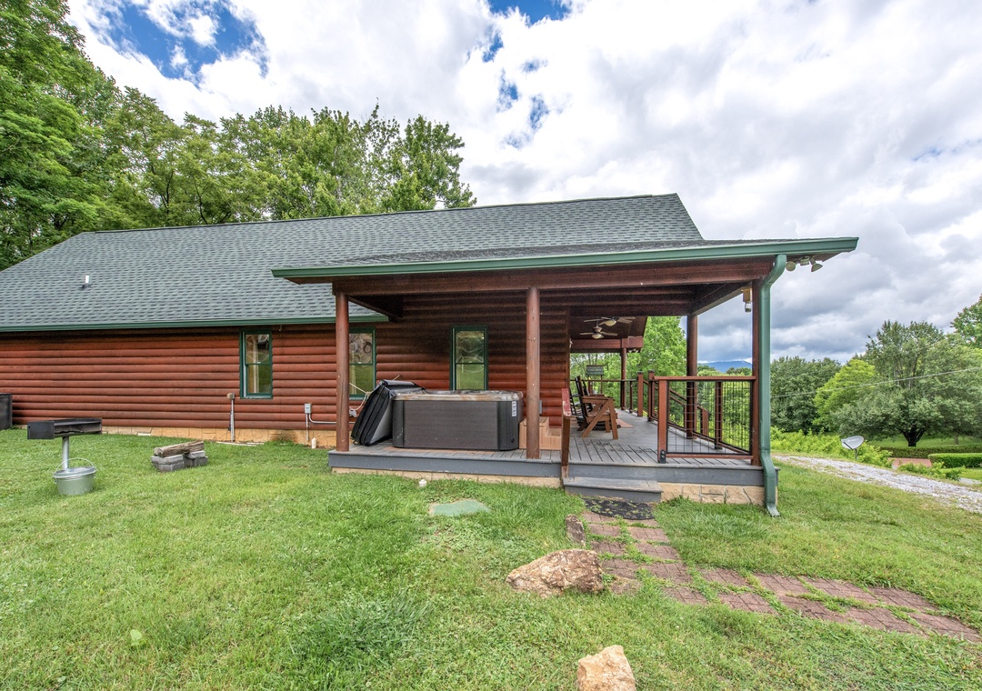 Charming log cabin exterior features a covered deck with hot tub, rocking chairs, and green metal roof, surrounded by lush lawn and mature trees in a peaceful countryside setting.