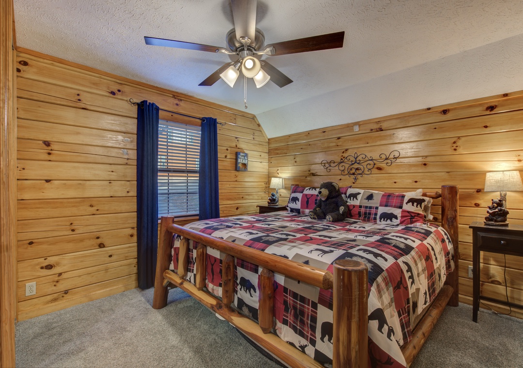 Unwind in your rustic mountain bedroom featuring warm wood walls, charming bear-themed bedding, and a ceiling fan for perfect comfort.