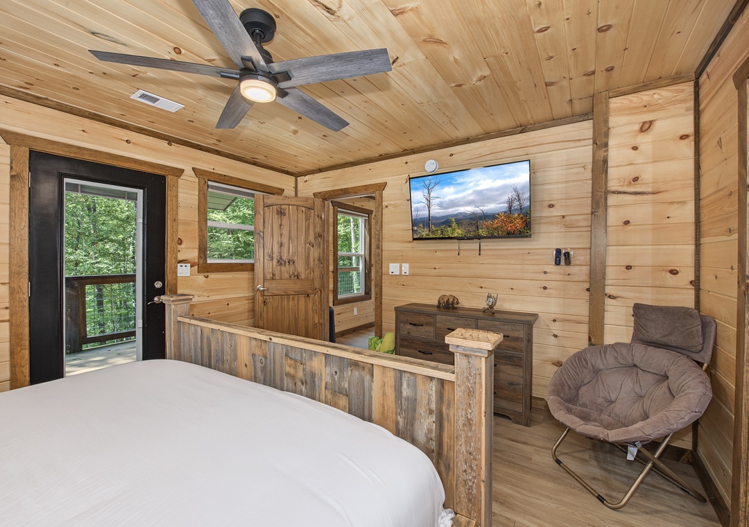 Unwind in your cozy cabin bedroom with warm wood walls, a ceiling fan overhead, and doors opening to your private deck. A comfy chair and rustic dresser complete the peaceful retreat.