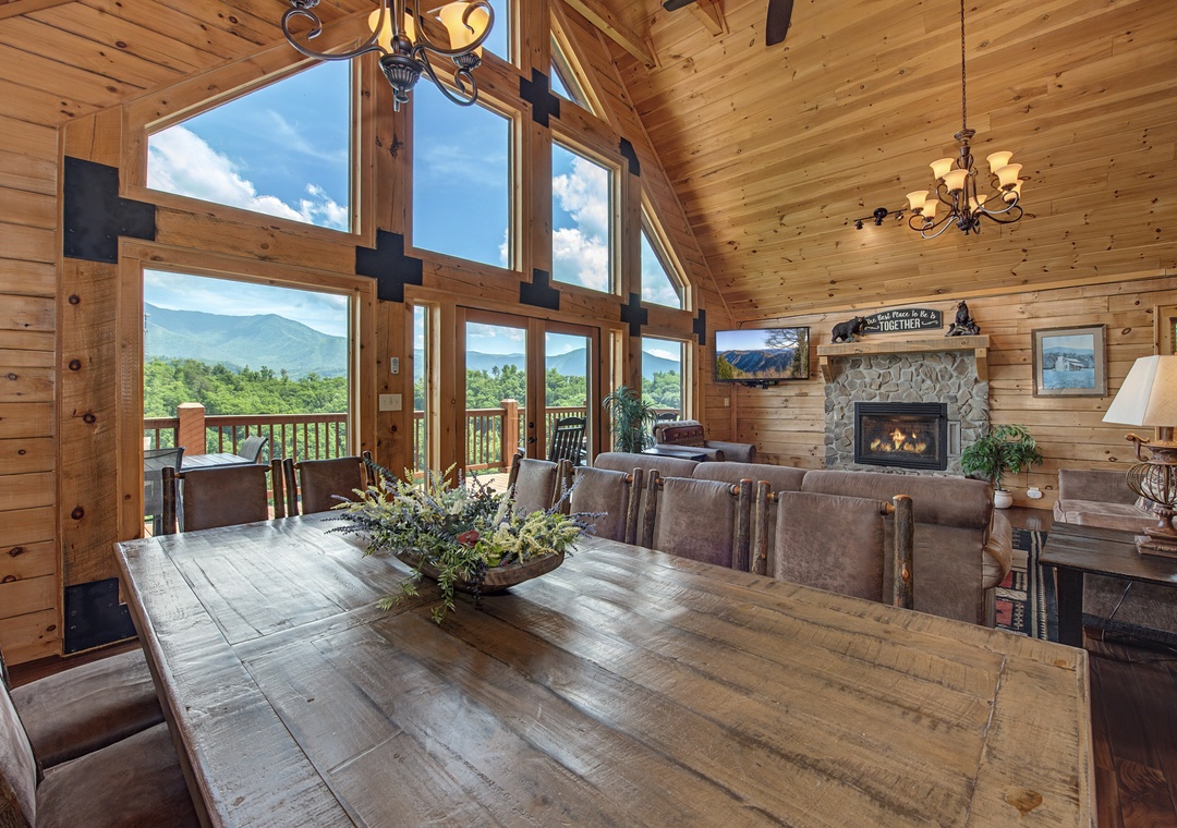 Gather around this expansive dining table where you can savor meals while soaking in breathtaking mountain views through floor-to-ceiling windows, with a cozy stone fireplace adding warmth nearby.