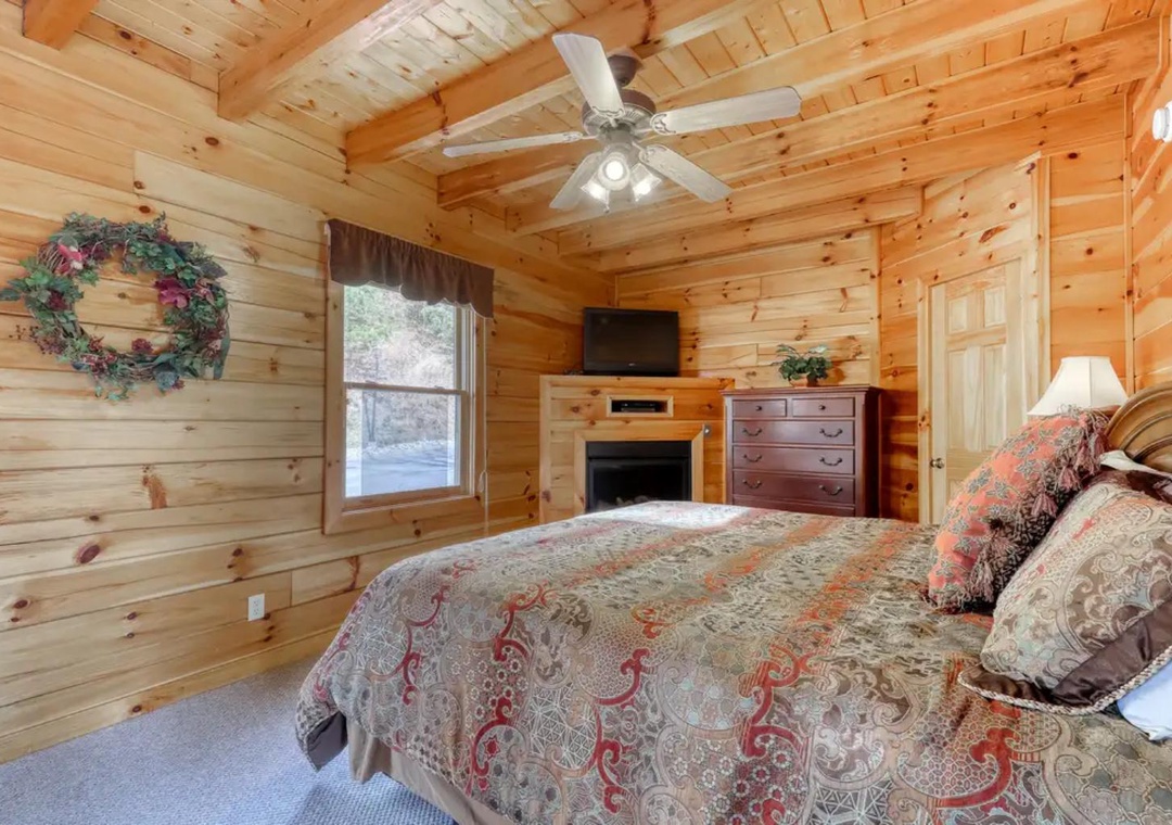 Unwind in your cozy cabin bedroom featuring warm wood walls, a crackling fireplace, and rustic charm. Natural light streams through the window as you relax under soft bedding with a ceiling fan overhead.