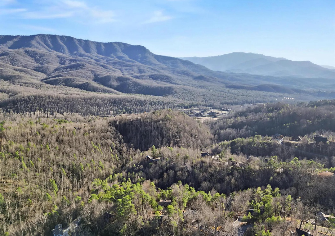 Aerial view showcasing the property nestled among wooded hills with dramatic mountain ridges rising in the distance under clear skies.