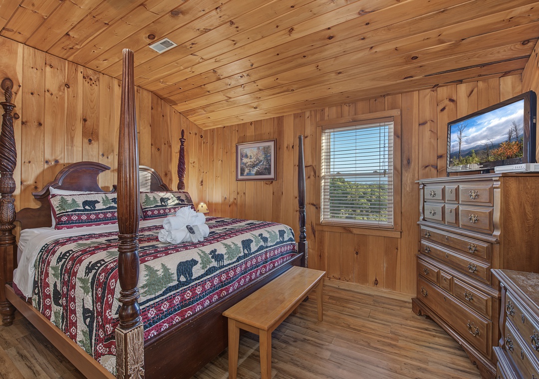 Your cozy cabin bedroom features a handsome four-poster bed with rustic charm, warm knotty pine walls, and a window framing serene greenery outside. Everything you need to unwind in mountain comfort.