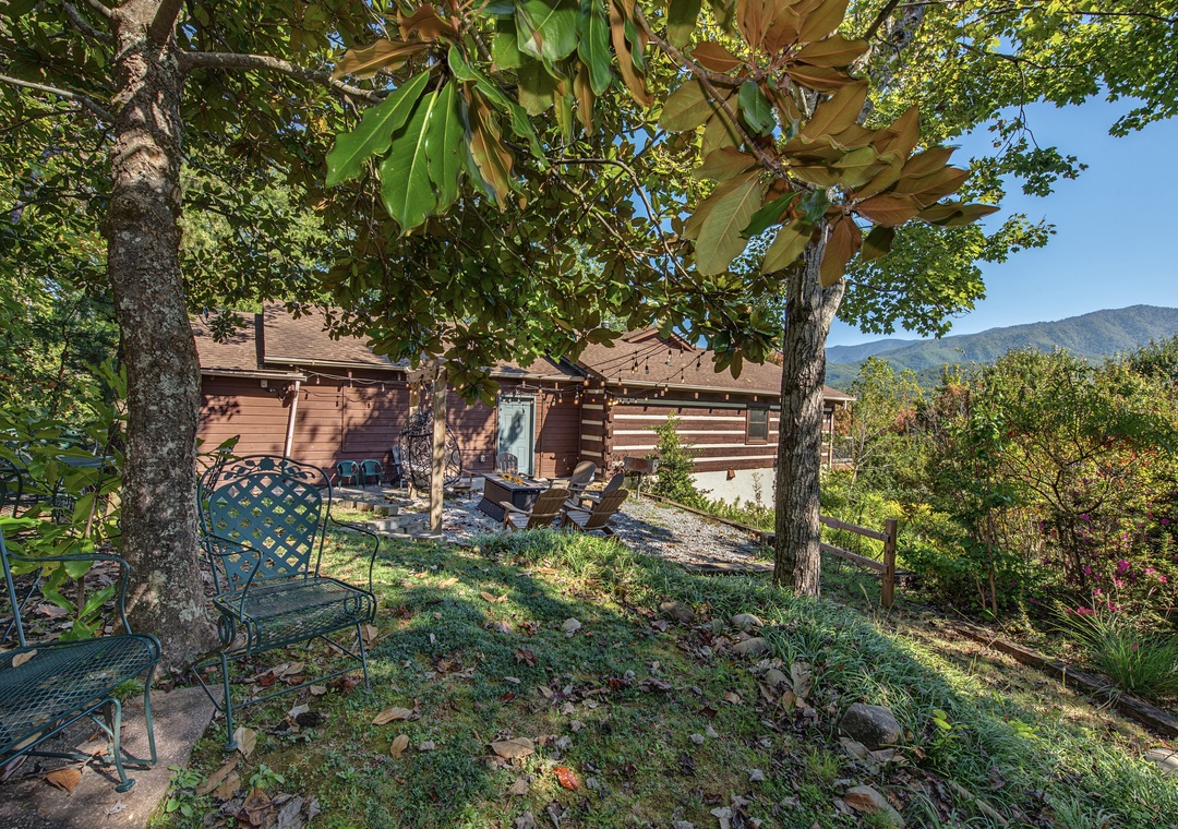 A charming cabin nestled beneath mature shade trees with mountain views visible in the distance. The outdoor seating provides a quiet spot to enjoy the natural landscape and surrounding greenery.