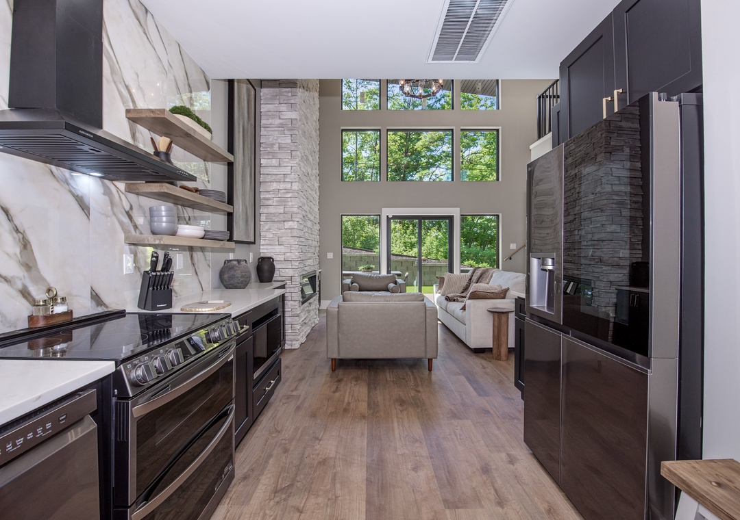 Stunning open-concept kitchen and living space with soaring ceilings and floor-to-ceiling windows framing lush forest views. Your modern chef's kitchen features sleek dark cabinetry, marble backsplash, and stainless appliances—perfect for preparing meals 