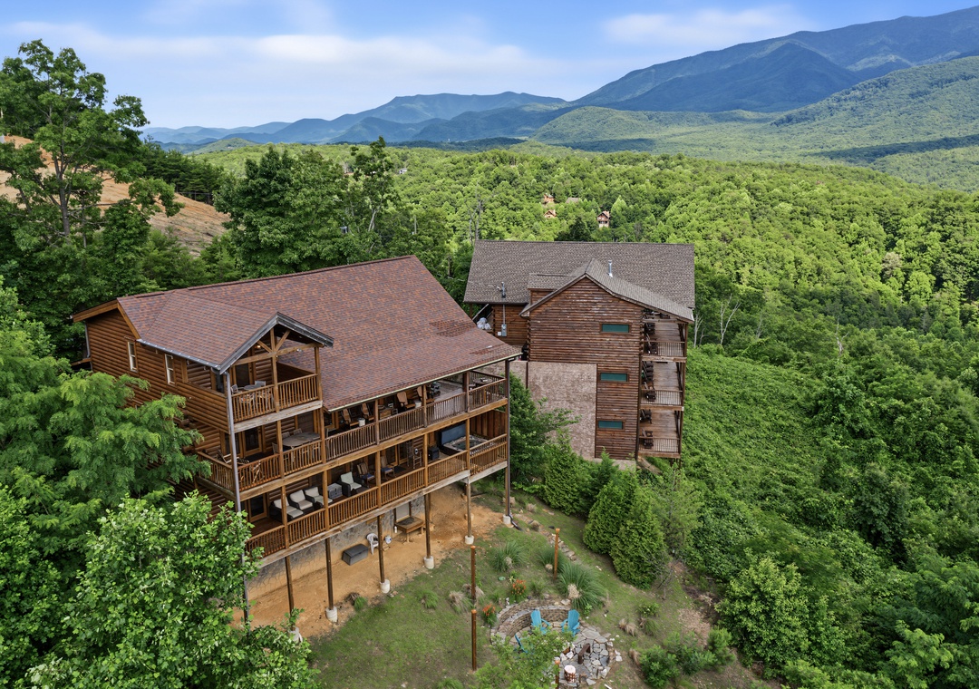 Elevated mountain chalets nestled in lush forest with sweeping views of layered mountain ridges and valleys beyond. The property is set amid abundant natural landscape and trees.