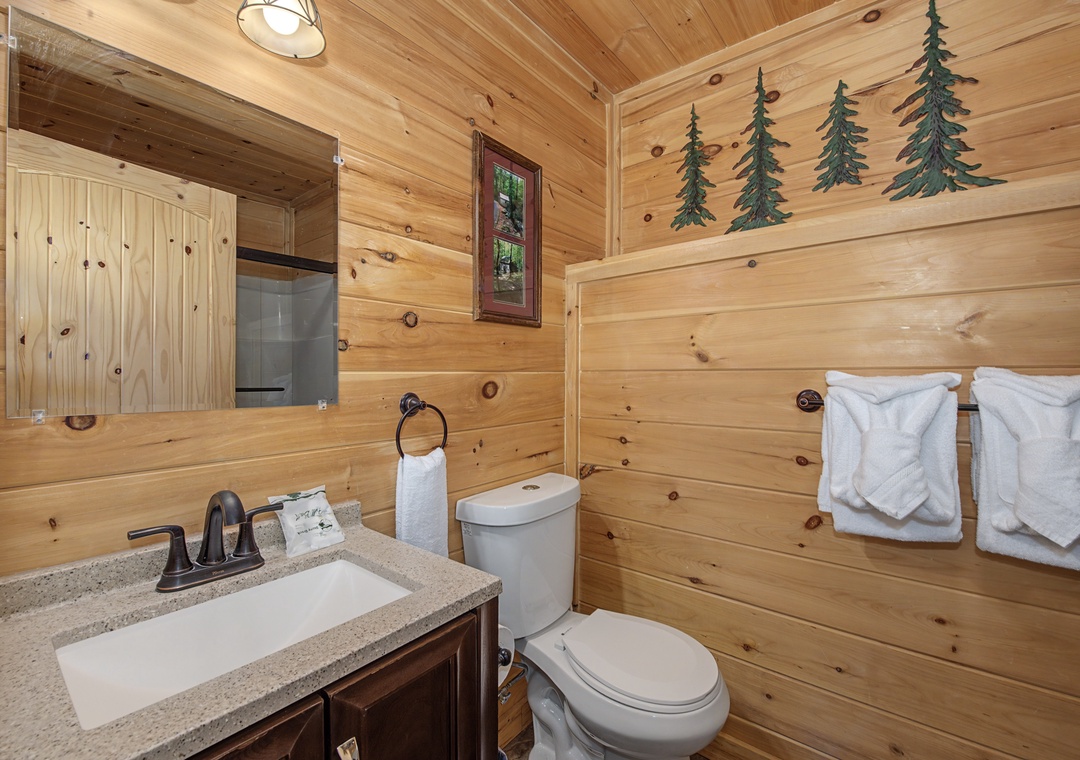 Freshen up in your cozy cabin-style bathroom, featuring warm pine walls adorned with rustic tree accents, plush white towels, and a charming woodland window view for a true mountain escape.