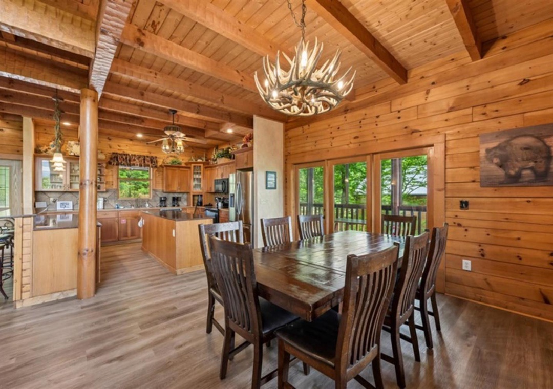 Gather around the rustic wood dining table under a striking antler chandelier. Natural light pours through large windows showcasing lush greenery, while the open kitchen invites easy entertaining in this warm, cabin-style retreat.