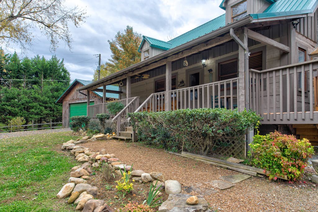 Rustic cabin exterior with a distinctive teal metal roof, wraparound wood deck, and landscaped grounds with stone pathways and mature evergreens. The property blends mountain charm with natural surroundings.