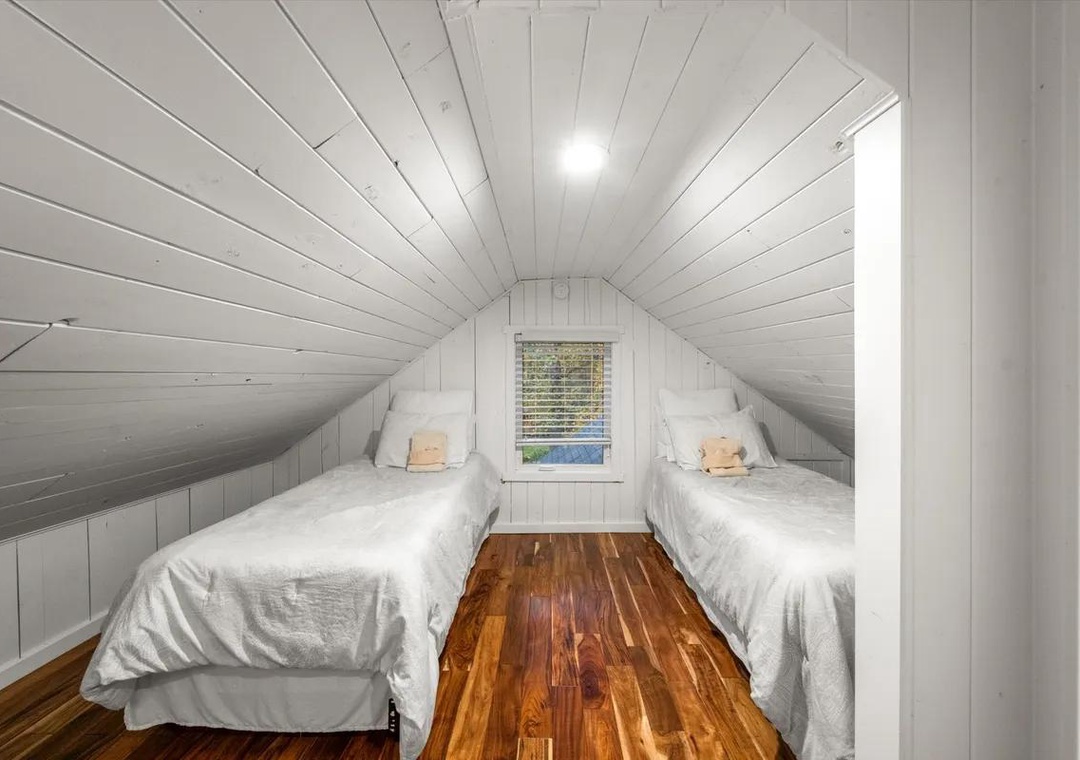 This cozy attic bedroom features two twin beds with plush white linens, warm hardwood floors, and charming whitewashed shiplap walls and ceiling—perfect for peaceful rest after a day of adventure.