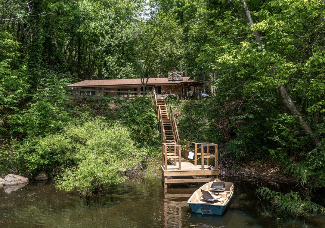 This serene riverside retreat features a private wooden dock and boat, offering direct water access. The property sits nestled among towering trees, connected by a charming staircase leading to the water's edge.