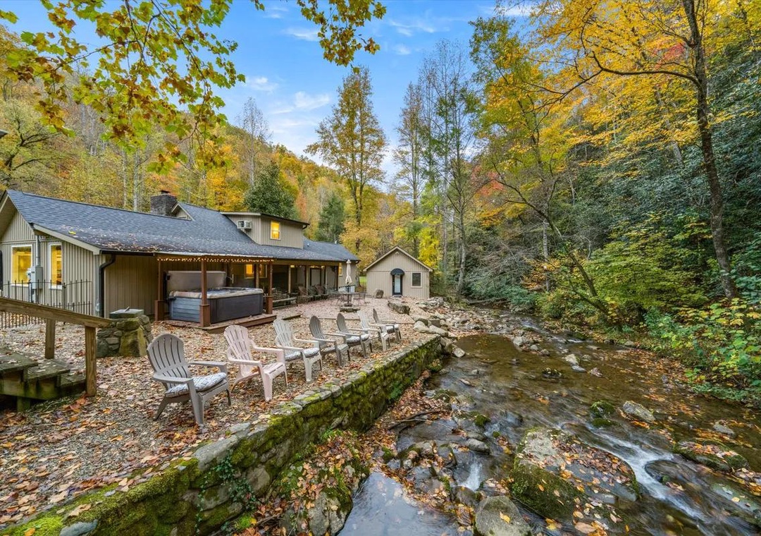 This autumn retreat nestled along a cascading creek showcases peaceful outdoor living areas, with a spacious patio, covered hot tub pavilion, and cozy Adirondack seating surrounded by vibrant fall foliage and mountain scenery.