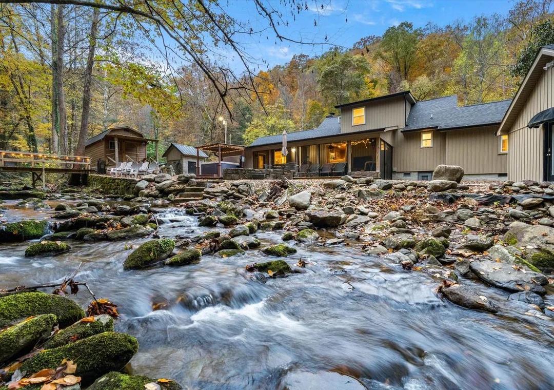 This property sits beside a gentle mountain stream with moss-covered stones, surrounded by autumn foliage and forested hills under a bright blue sky.