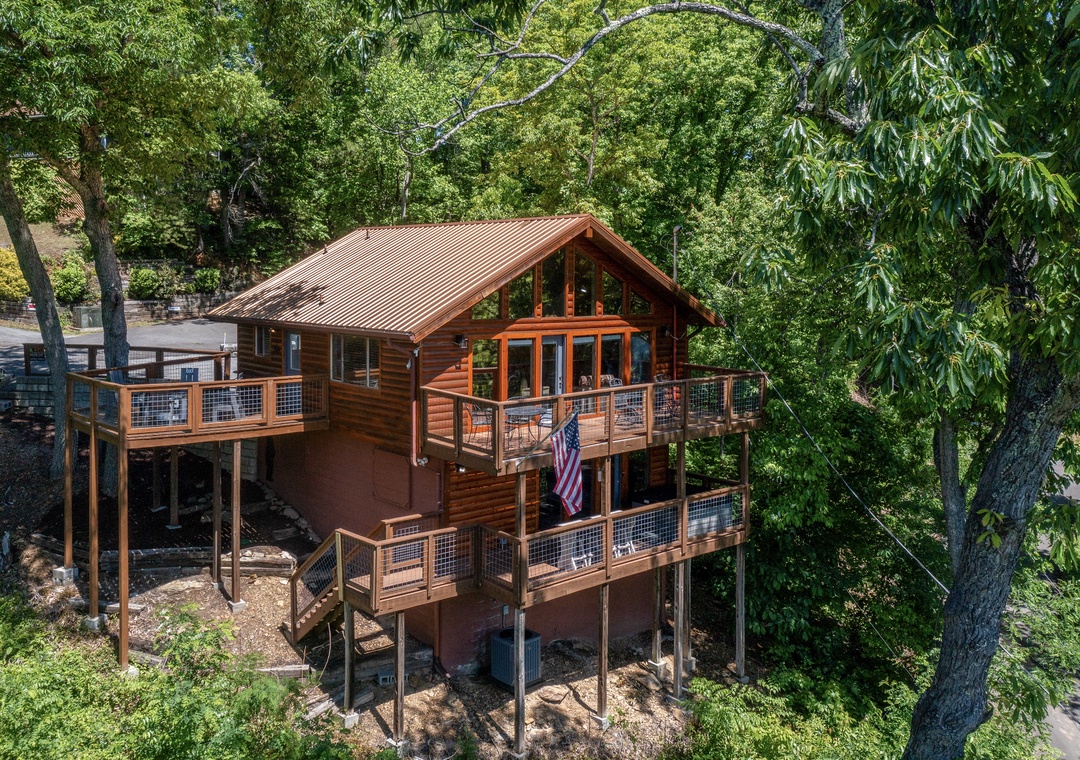 A charming hillside cabin nestled among towering trees, featuring multi-level decks and floor-to-ceiling windows. The property offers a secluded wooded setting with natural shade and rustic architectural details.