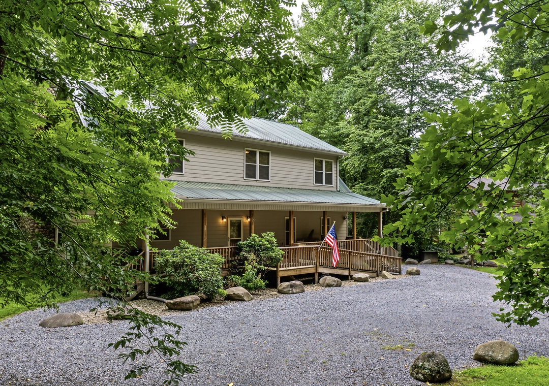 Charming two-story retreat nestled in lush greenery, featuring a welcoming wraparound porch and gravel driveway—a peaceful mountain escape surrounded by nature.