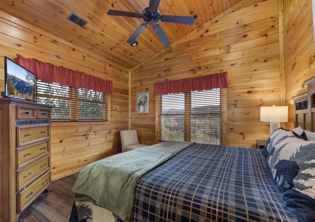 Curl up in your mountain retreat with knotty pine walls and a ceiling fan overhead. Natural light streams through dual windows, while plaid bedding and rustic wood furniture complete the cozy cabin feel.
