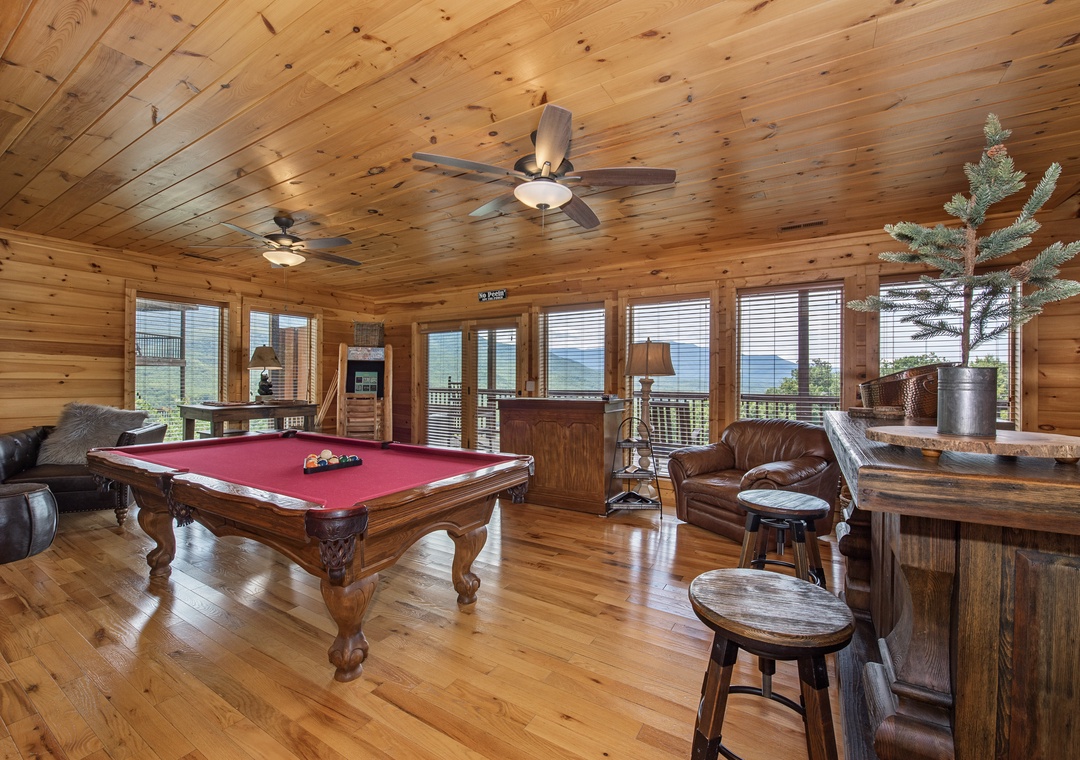 Gather with friends in your spacious game room, where a pool table and rustic bar await beneath soaring wood-paneled ceilings. Large windows frame sweeping mountain vistas, creating a warm, inviting space for unforgettable moments.