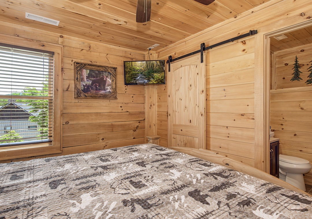 Relax in your cozy wood-paneled bedroom featuring rustic barn doors, a plush bed, and a wall-mounted TV. Soothing nature views and cabin charm await.