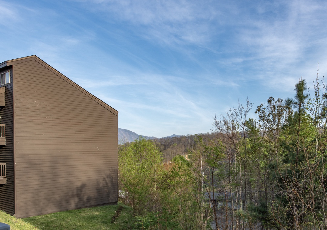 Multi-story brown wooden building nestled among rolling mountains and lush spring greenery with distant peaks visible.