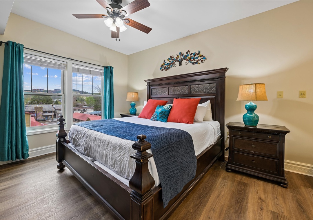 Your elegant bedroom features a plush king bed with vibrant linens, matching teal lamps, and cheerful windows overlooking the neighborhood.