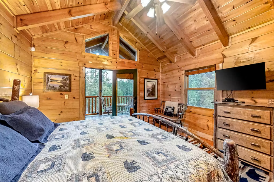 Your rustic log cabin bedroom features warm wood walls, cozy quilted bedding, and peaceful forest views from your private balcony.