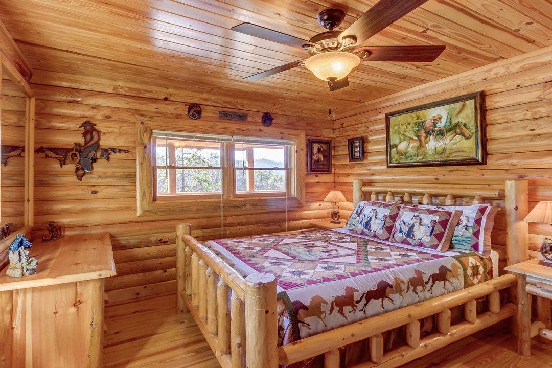 Your cozy Western-themed bedroom features a handcrafted log bed and warm wood paneling throughout. The ceiling fan keeps the space comfortable, while natural light floods through double windows overlooking the surrounding trees.