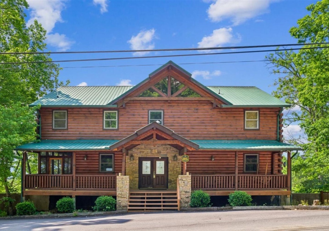 This expansive log cabin-style building features a striking stone and timber entrance, twin covered porches, and a distinctive green metal roof—set in a lush wooded area for a peaceful mountain retreat.