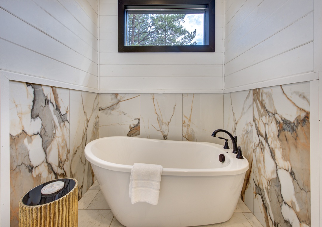 Unwind in your elegant freestanding soaking tub, surrounded by stunning marble walls and natural light from the tree-lined window above.