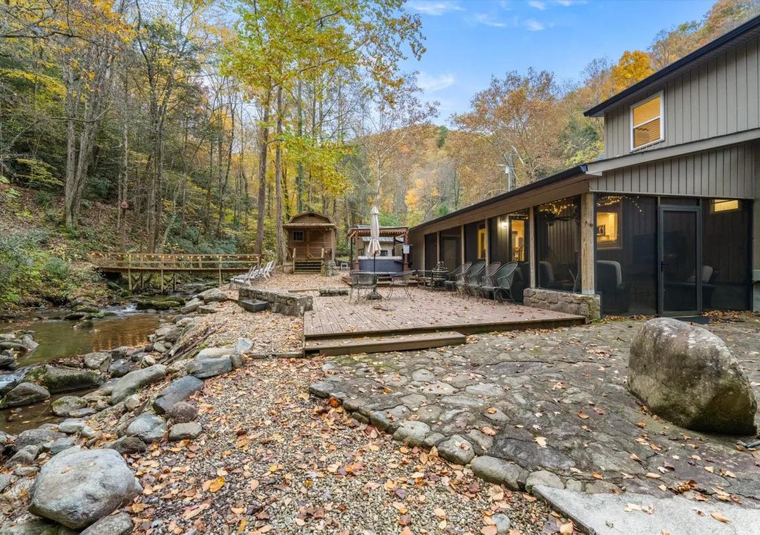 The property nestles beside a gentle creek in a wooded mountain setting, with a stone patio, hot tub, and covered deck creating a private retreat surrounded by autumn foliage.