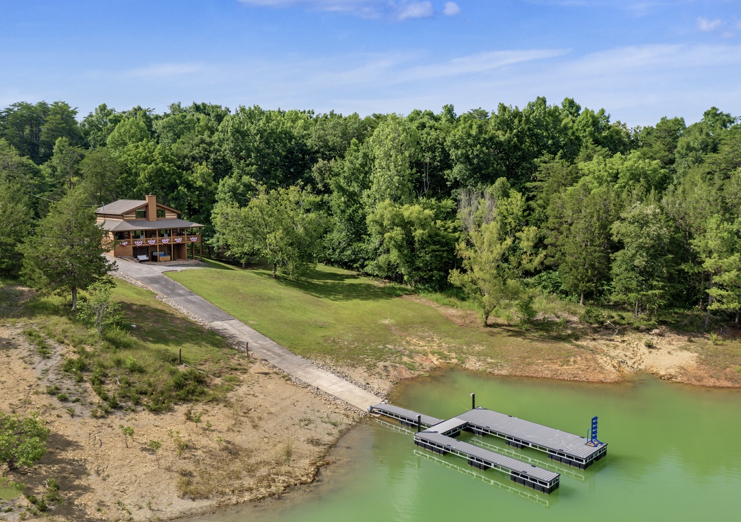 Aerial view of a lakeside vacation rental property featuring a private dock, expansive lawn, and serene natural surroundings with lush forest backdrop.