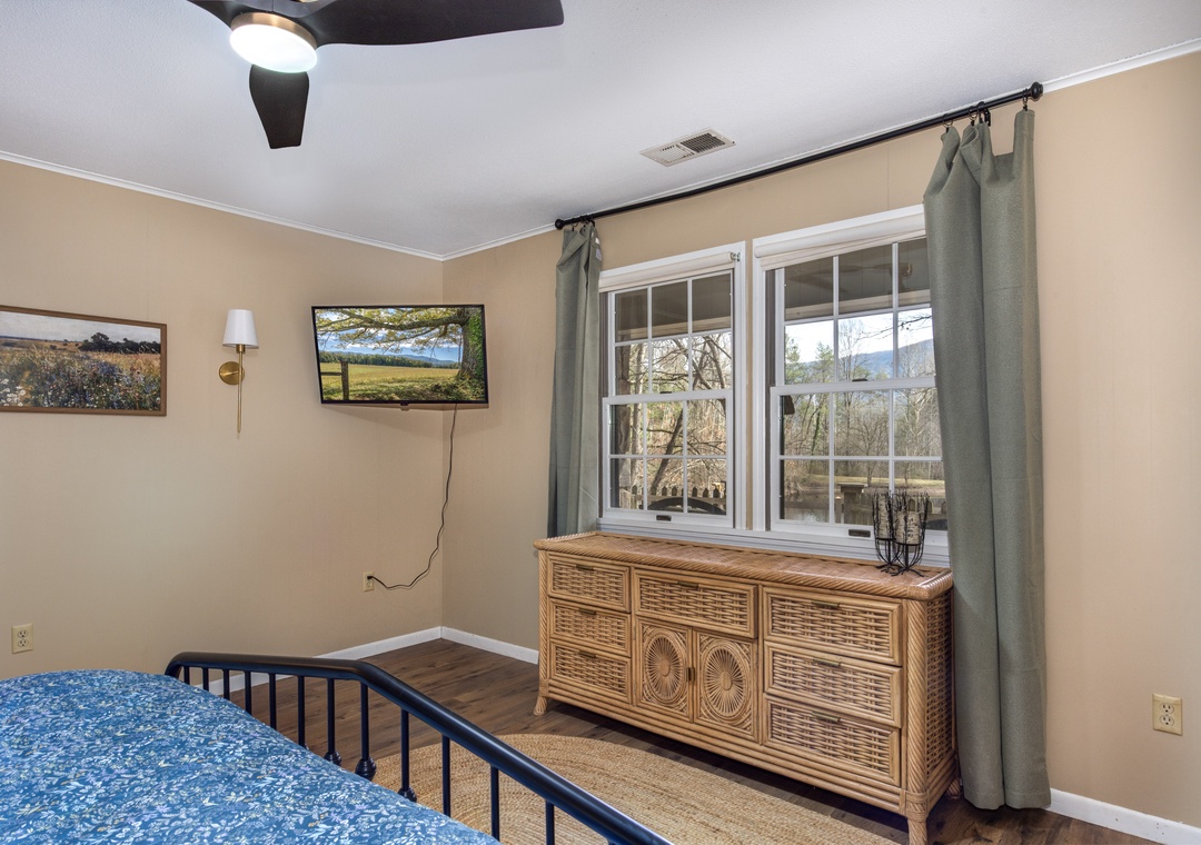 Your peaceful bedroom features a ceiling fan, wall-mounted TV, and natural light streaming through large windows overlooking scenic views.